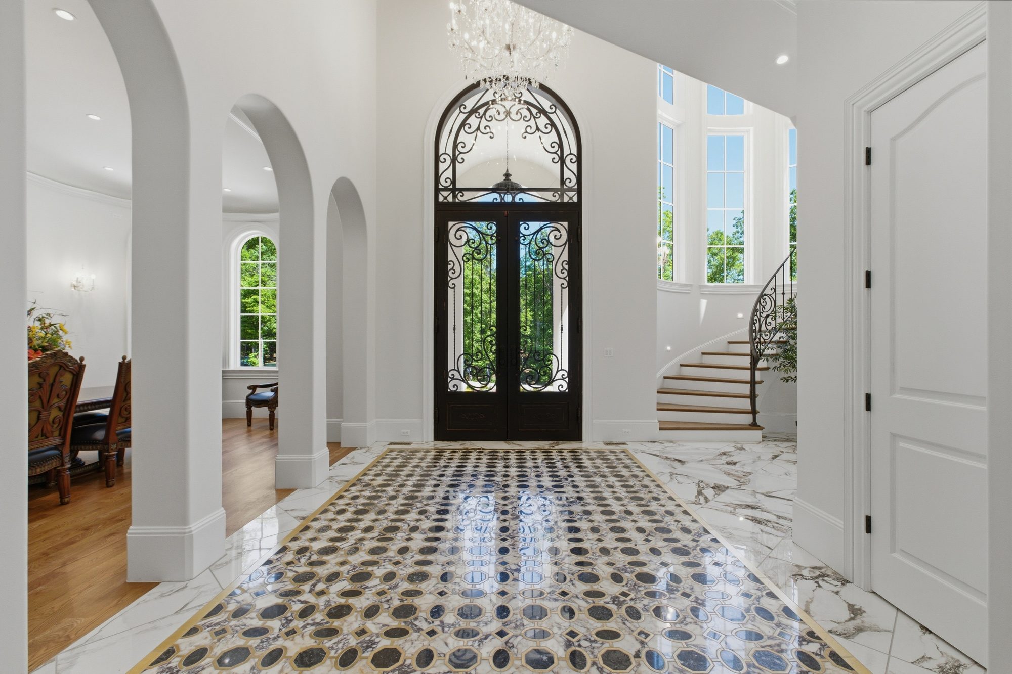 The two-story foyer with vaulted ceiling, ornate iron-and-glass arched doors, crystal chandelier, marble and brass inlay floor, arched colonnades, and a curved staircase
