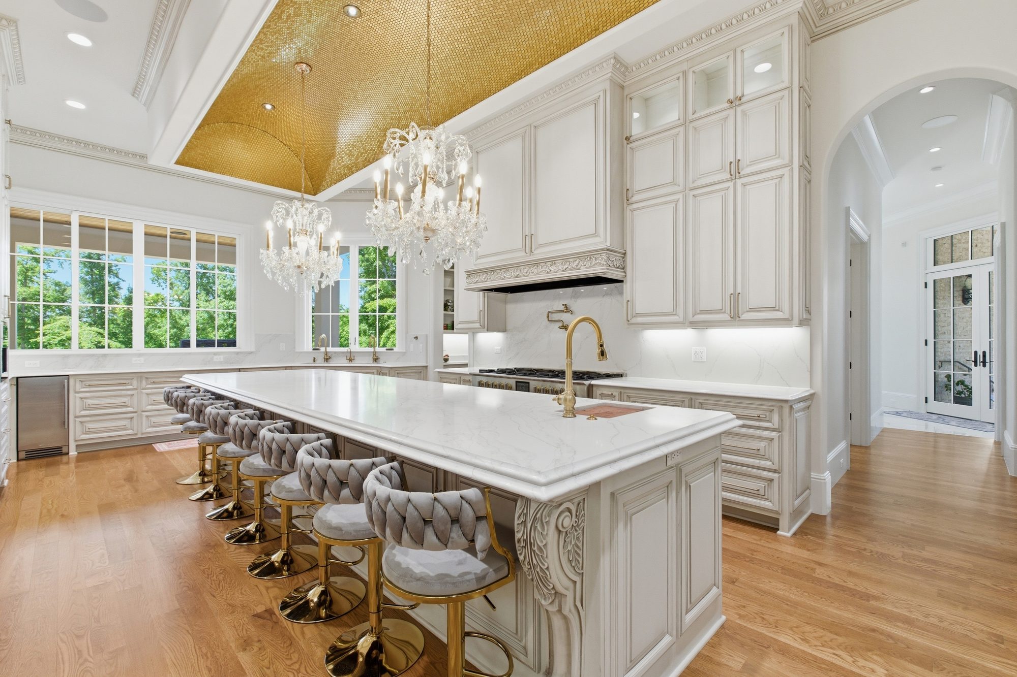Three-quarter view of the chef's kitchen showing the marble island, range with marble backsplash, and tall windows opening to the trees