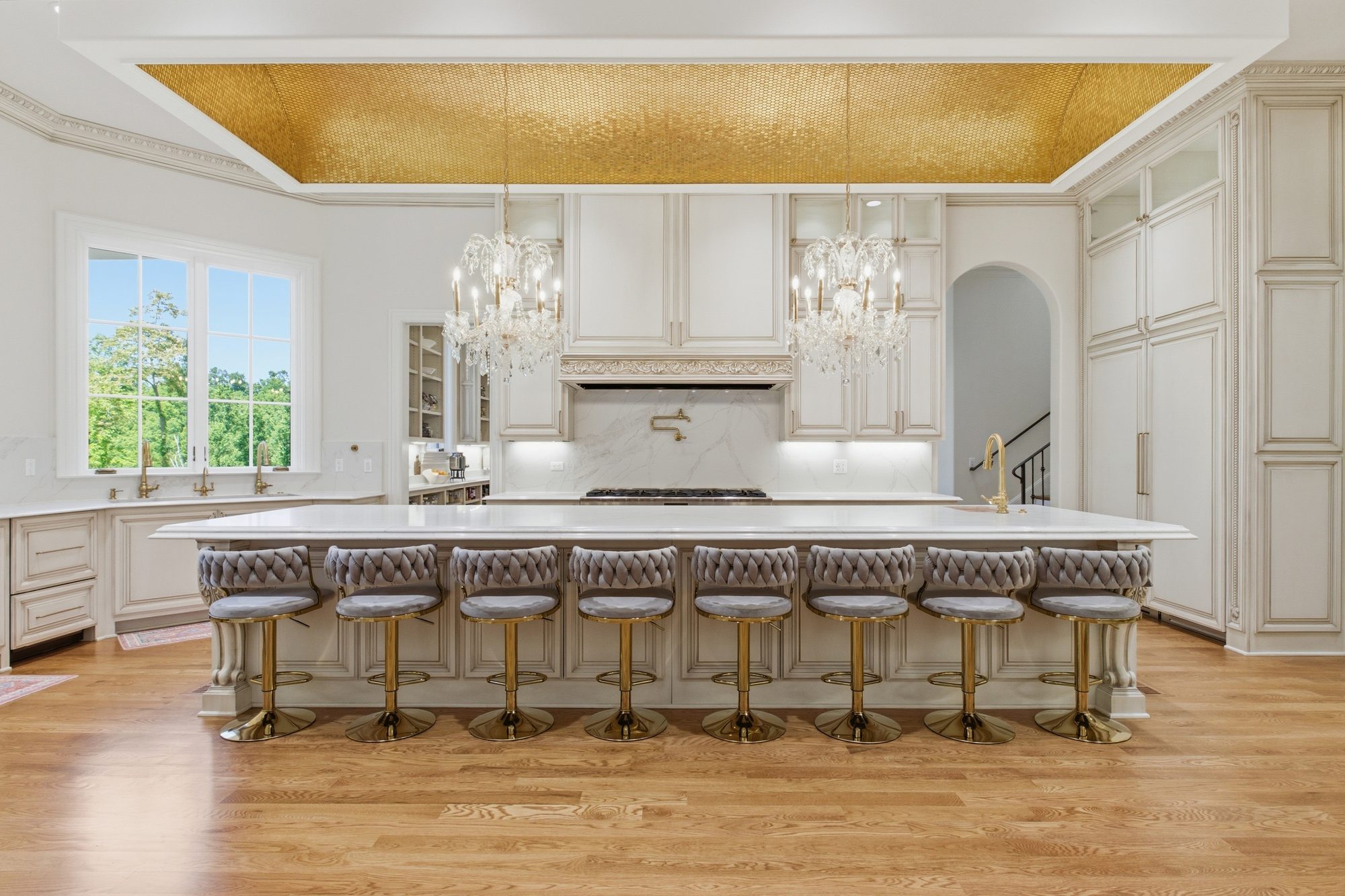 The chef's kitchen at full view — gold mosaic barrel-vault ceiling, twin crystal chandeliers, marble island, and a long run of velvet bar stools with gold bases