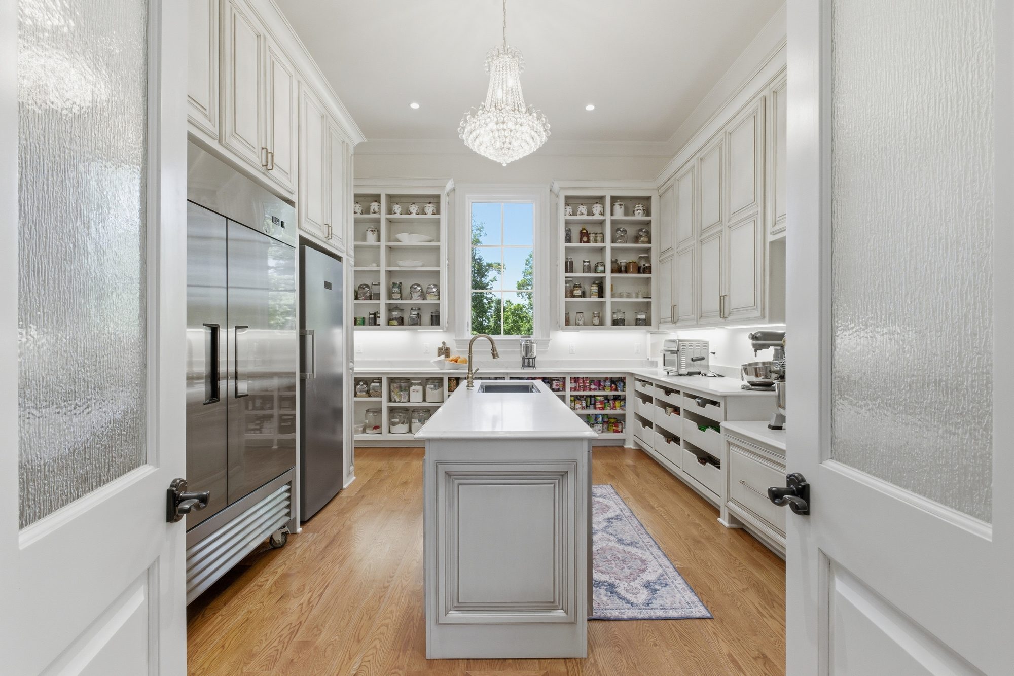 Working scullery beyond the chef's kitchen with secondary island, full-height refrigeration, open shelving, and crystal chandelier
