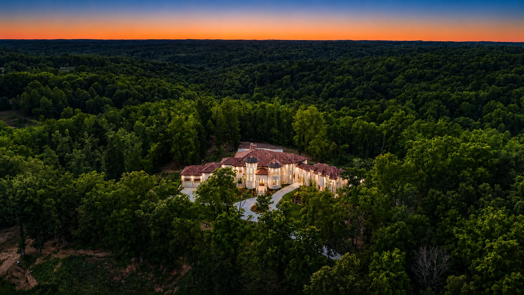 Aerial view of the estate set deep within wooded hills at sunset, the home glowing against an endless canopy of hardwoods