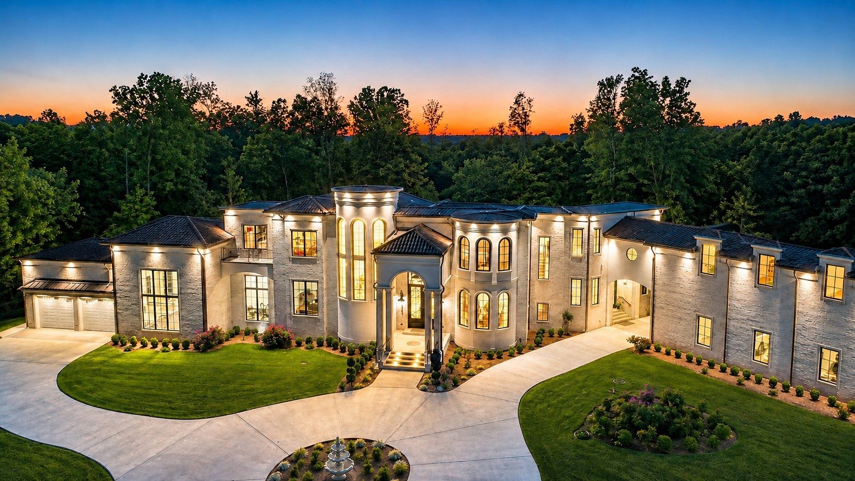 The estate at twilight, windows aglow against a warm sunset sky and surrounding hardwoods